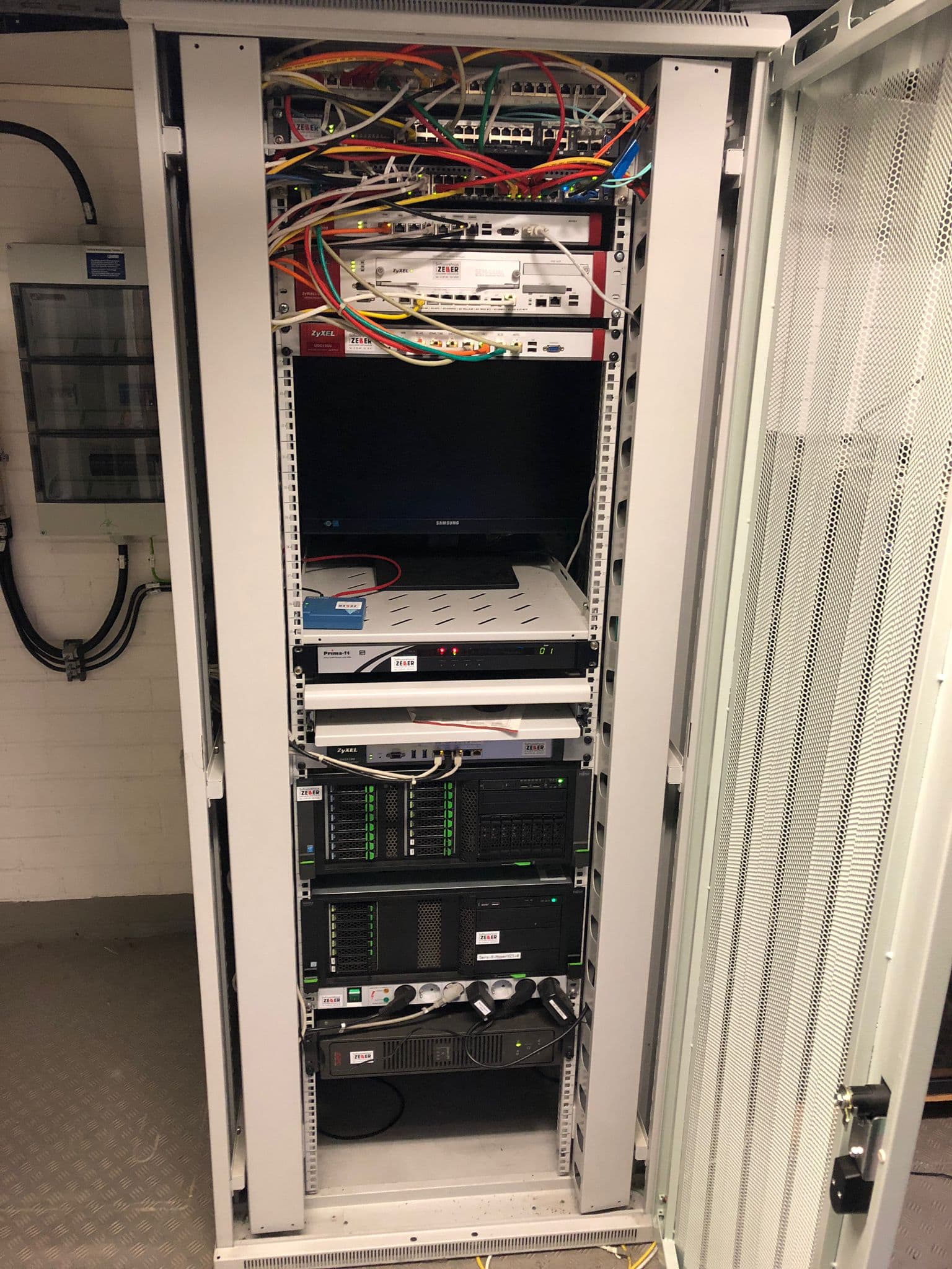 Weißer Serverschrank mit bunten Kabeln, Zyxel-Routern, einem Samsung-Monitor und mehreren Servern in einem Technikraum.