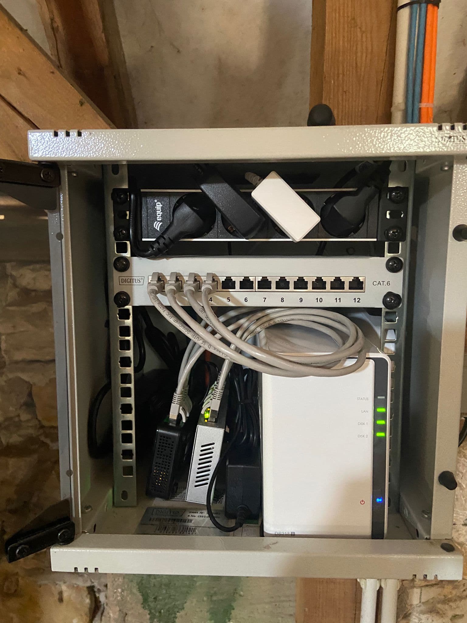 Kompakter Netzwerkschrank mit Patchpanel, weißem Synology NAS und Stromleiste vor einer rustikalen Steinwand.